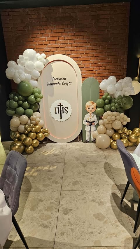 First Communion altar decoration featuring an arched white backdrop with IHS cross symbol, surrounded by white, green, and gold balloons, with a child figurine in white communion attire.