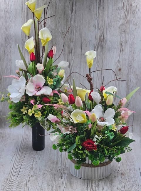 Two spring floral arrangements with yellow calla lilies, pink flowers, red tulips, and greenery in black and white vases on weathered wood surface