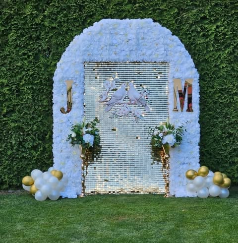 Decorative event backdrop with blue-white flower arch, shiny metallic door, gold and white balloons on grass