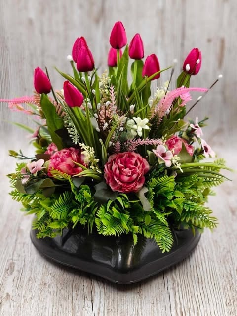 Pink tulips and peonies in a black vase with ferns and white flowers on a light wooden background