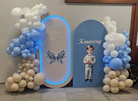 Decorative arch display with blue neon-lit panels, white and blue balloons, and text reading Pierwsza Komunia Swietla and Krawery