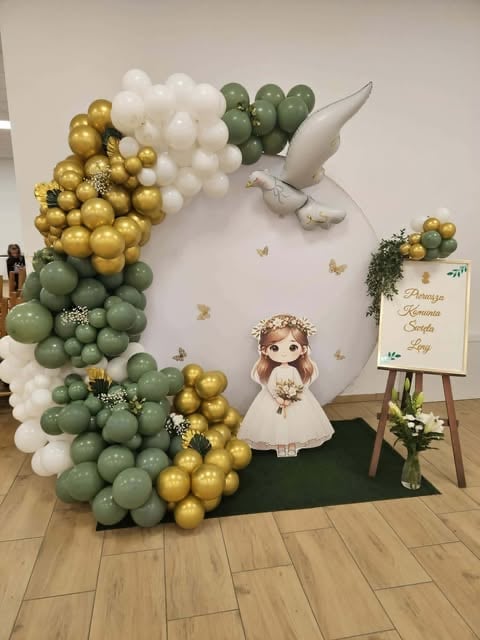 Party backdrop with balloon garland in green, gold, and white, featuring a white dove, decorative girl figure, and welcome sign