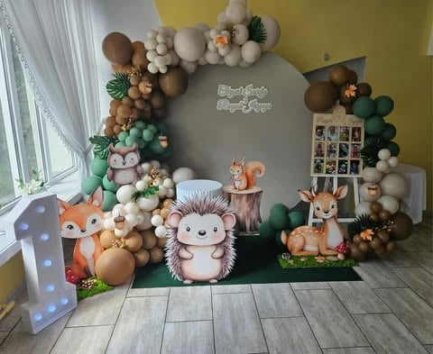 Woodland-themed party backdrop with forest animal cutouts, colorful balloon garland, and photo display on gray wall.