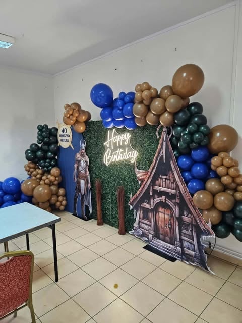 Birthday party setup with blue, brown, and dark green balloon arch, Happy Birthday neon sign on moss wall, and Halloween-themed haunted house cardboard cutout